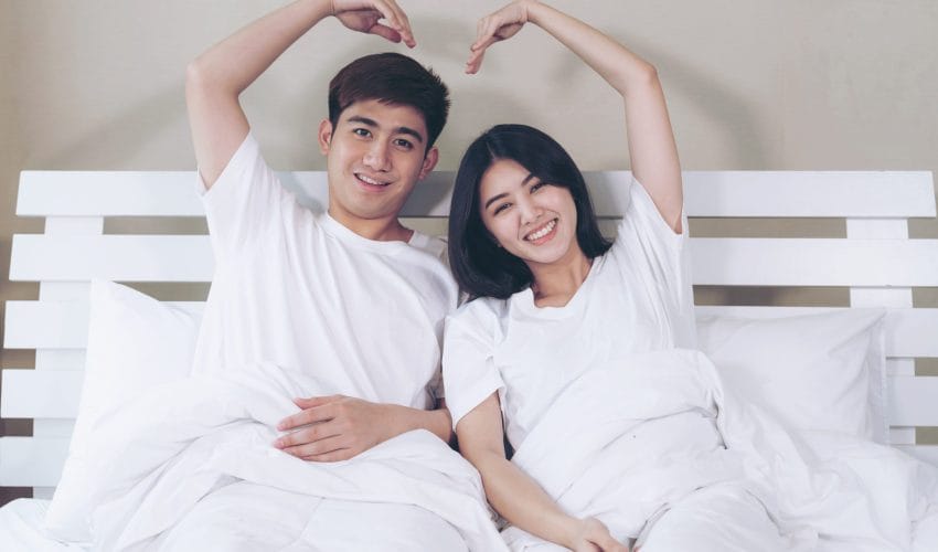 Portrait happy young couple sitting and smiling on bed and raise up arms make heart shape together
